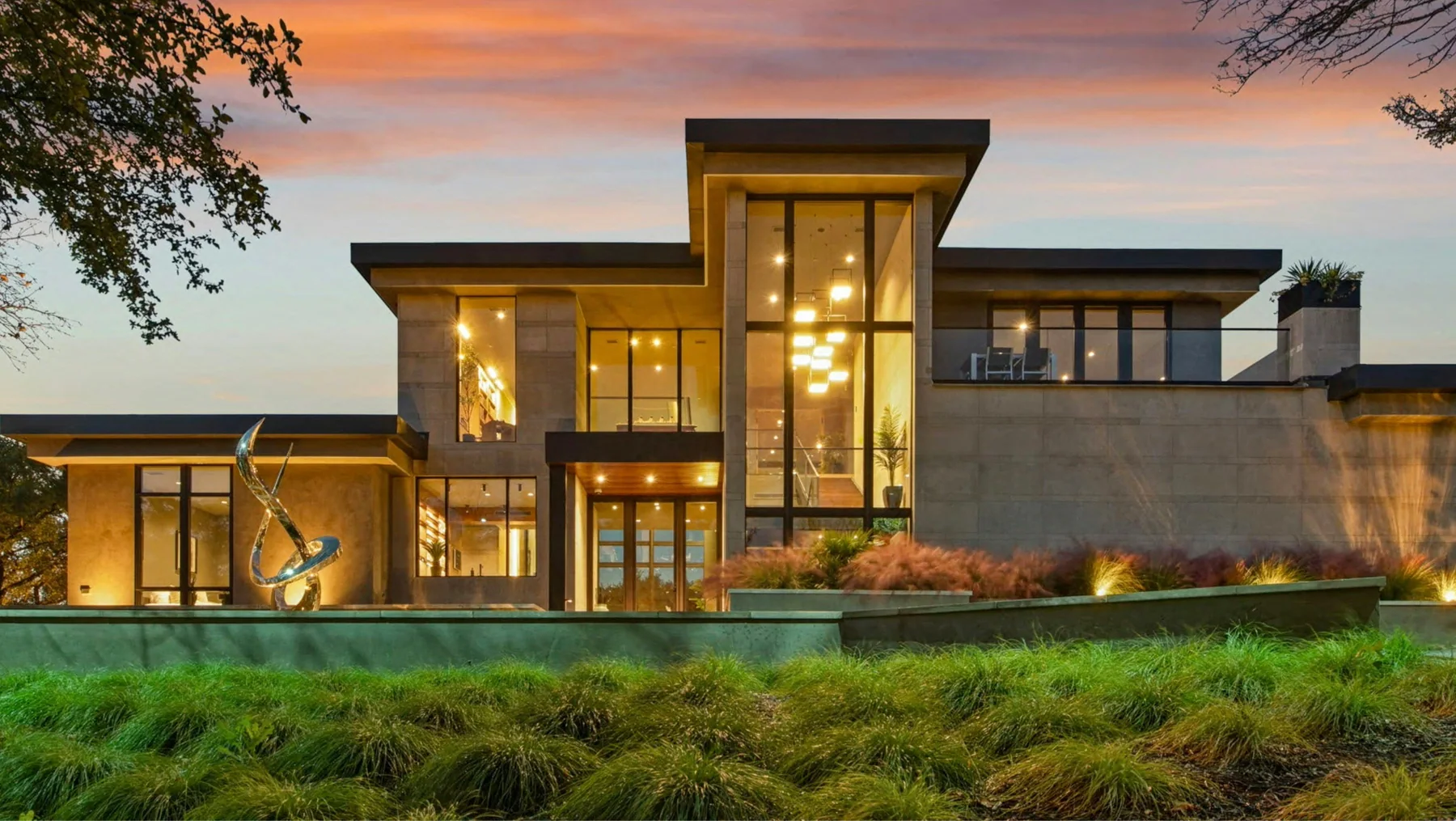 A modern glass-and-stone home at dusk with warm light in every window